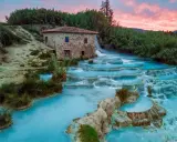 Cascate termali turchesi in Toscana, con un antico mulino in pietra e un cielo rosa al tramonto. Curiosità sulla Toscana: un luogo magico.