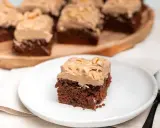 En bit chokladtårta med krämig frosting och nötter. Flera bitar serveras på ett träfat i bakgrunden.