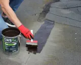 Naprawa przeciekającego dachu z papy. Rękawica w czerwonej rękawicy nakłada ciemną masę uszczelniającą na pęknięcia.