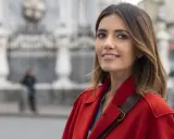 Donna con cappotto rosso sorride, con uno sfondo sfocato di edifici e una persona. Forse sta pensando ai **mina settembre libri** da leggere.