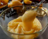 Cuillère prélevant une généreuse portion de crème pâtissière citron, onctueuse et dorée, dans un bol en verre. Des citrons flous en arrière-plan.