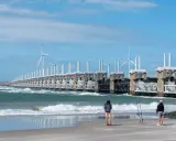 Dwie osoby stoją na plaży, obserwując falujące morze i imponującą zaporę z turbinami wiatrowymi. Takie morze jest w Holandii, chronione przez innowacyjne konstrukcje.
