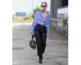 Donna con camicia a righe blu e bianche, occhiali da sole e un elegante outfit pantaloni ecopelle neri con zip frontale. Porta una borsa verde oliva.