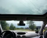 Widok z wnętrza samochodu z panoramicznym dachem, przez który widać niebo i drzewa. Krople deszczu na szybie.