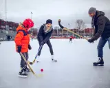 Familj njuter av ishockey i Göteborg. En pappa, mamma och son med hockeyklubbor och en röd puck på isen.