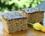 Zwei Stücke saftiger Zucchini Kuchen mit Schokoladenglasur und gehackten Pistazien, daneben eine Zucchiniblüte.