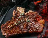 Un jugoso chuleton a la brasa, con romero fresco, listo para ser servido junto a brasas ardientes.
