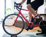 Intensywny trening na trenażerze kolarskim w domu. Czerwony rower, sportowiec w spodenkach i koszulce, butelki z wodą.