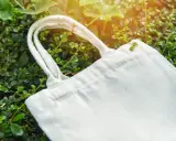 Eine weiße Stofftasche liegt zwischen grünen Blättern. Perfekt, um die Einkaufsstaschen zu verstauen und die Umwelt zu schonen.