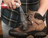 Jak zabezpieczyć nowe buty skórzane? Mężczyzna spryskuje brązowe buty trekkingowe preparatem ochronnym.