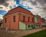 Tykocin co zobaczyć: zabytkowa Wielka Synagoga z różową fasadą i zielonym aneksem, na brukowanym placu pod burzliwym niebem.
