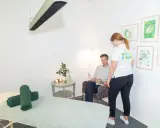 En kvinna i vit tröja med texten "HÄLSO RESURS" pratar med en man som sitter i en fåtölj. Rummet har en massagebänk och konst på väggarna, kanske för att förbereda för en vagusnerven massage.