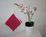 Två tjocka, virkade grytlappar i rosa med vit kant hänger på en vit vägg bredvid en orkidé i kruka.