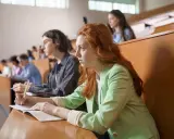Studentka z rudymi włosami skupiona na wykładzie, robi notatki. Jej kolega obok również studiuje psychologię.