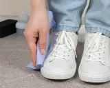Persona limpiando zapatillas blancas con un paño azul. A su lado, un cepillo y un bote de limpiador.