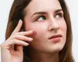 Une jeune femme aux yeux bleus regarde au loin, sa main effleurant sa joue. Ses cils, sublimés par un **tatouage cils**, accentuent son regard captivant.