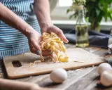 Hände halten frisch gemachte, italienische Pasta. Eier und Zutaten liegen bereit, um italienische Pasta selber machen zu können.