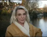 Pia Degermark idag, med sitt blonda hår och en varm fårskinnsjacka, står vid en vattensamling med träd och hus i bakgrunden.