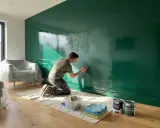 Hombre lijando pared verde brillante, preparándose para pintar. Muebles cubiertos, latas de pintura a mano.