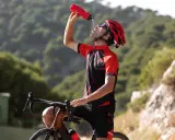 Kolarz w kasku i stroju sportowym pije wodę z bidonu, odpoczywając po intensywnym treningu, który podnosi VO2 max.