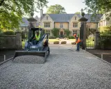 Un mini-escavatore scarica ghiaia per creare un vialetto giardino ghiaia davanti a una villa. Un operaio livella la ghiaia con un rastrello.