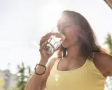 Kobieta w żółtej bluzce pije wodę, dbając o nawodnienie po alkoholu. Słońce przebija przez jej włosy.