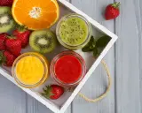 Bandeja con fresas, rodajas de kiwi, naranja y tres tarros de **puré de frutas** de colores vibrantes: verde, amarillo y rojo.