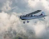 Eine braun-weiße Cessna 170 fliegt durch die Wolken über grünen Feldern.