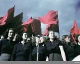 En grupp kvinnor deltar i ett 1 maj tåg, med röda fanor vajande i bakgrunden.