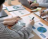 Des mains travaillent sur des documents financiers, une calculatrice et un ordinateur portable. L'ergonomie organisationnelle est au cœur de cette réunion axée sur le développement durable.