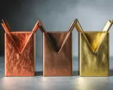 Trois porte-stylos aux formes géométriques, aux teintes cuivrées et laiton couleur, sur un fond gris.