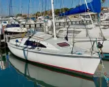 Un voilier blanc avec une voile bleue dans un port, reflété dans l'eau calme.