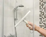 Hand mit Abzieher reinigt Duschkabine. Wasserflecken und Seifenschaum werden entfernt, um die Dusche zu putzen.