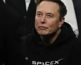Elon Musk w bluzie SpaceX, zamyślony, w otoczeniu innych osób.