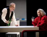 Krister Henriksson i en dramatisk scen. Han håller en grön flaska, medan en kvinna i rött med ett glas vatten ser på. Scenen påminner om hans roller i filmer och tv-program.