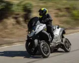 Un motorista con casco amarillo y negro conduce una moto de tres ruedas por una carretera. La moto ciudad se mueve rápido, creando un efecto de desenfoque.