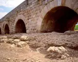 Antiguo puente de piedra con arcos, que es un puente de gran tamaño y robustez, con ruinas a sus pies.