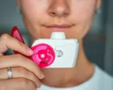 Nahaufnahme einer Person, die einen Pulverinhalator hält, bereit zur Anwendung. Der Inhalator ist weiß mit pinken Akzenten und zeigt die Dosis "27".