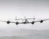 Ein vier-motoriger Propellerflieger mit markantem Leitwerk fliegt über bergiger Landschaft.