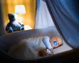 Bambino dorme sereno nella culla, con ciuccio, sotto una coperta calda. Un orsacchiotto veglia, simbolo di addormentamento autonomo.