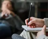 Psycholog pomaga zrozumieć siebie i swoje problemy. Na zdjęciu widać rękę terapeuty piszącą w notatniku, podczas gdy pacjent leży na kanapie.