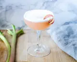 En rosa rabarber sour-drink med skum och en rabarberdekoration. Två stjälkar rabarber ligger bredvid.
