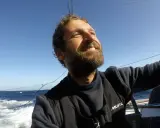 Un homme de mer souriant, le visage tourné vers le ciel bleu, navigue sur un voilier. L'océan agité et le soleil radieux témoignent de son aventure.