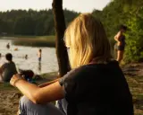 Dziewczyna z blond włosami siedzi nad jeziorem Trzechowskim, obserwując ludzi kąpiących się w wodzie.