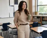 A woman in a tan sweater and slacks, holding a green tumbler, embodies how to dress like a teacher with a polished yet comfortable classroom look.