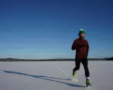 Mann mit grüner Mütze läuft im Winter über eine verschneite Fläche, die Sonne wirft lange Schatten.