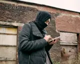 Osoba w czarnej chuście i kurtce trzyma telefon na tle zrujnowanej ceglanej ściany.