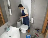 Instalator montuje WC kompakt z odpływem pionowym. W łazience widać umywalkę, lustro i narzędzia.