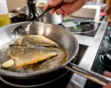 Un chef cocina pescado en una sartén de acero inoxidable, bañándolo con aceite caliente.