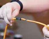 Ręce trzymające pomarańczowy kabel, który jest uszkodzony. Pokazuje, jak połączyć kable bez lutowania, używając taśmy izolacyjnej.